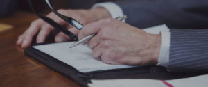 close up a lawyer's hands holding a pen and glasses and resting his hands on a notebook on a conference table